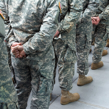 Photo: people in army fatigues lined up single file looking at their backs with the hands clasped behind them.