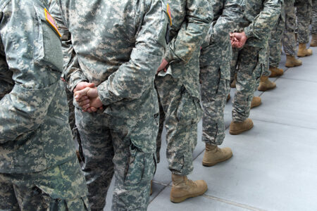 Photo: people in army fatigues lined up single file looking at their backs with the hands clasped behind them.