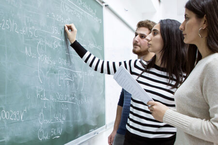 Photo of three young students writing a formula on a green blackboard with white chalk.
