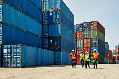 Photo: stacked shipping containers with a few worked in hard hats walking along side.