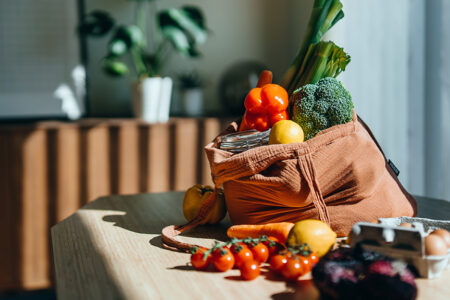 Photo of groceries including peppers, broccoli, lemons and eggs, on a kitchen table in a reusable shopping bag.