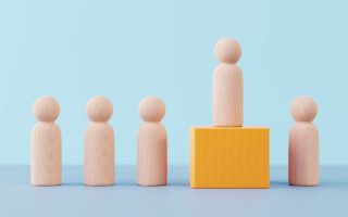 Photo: Wooden figurine pawn standing on wooden cube above other wooden figurines against minimal blue background. The concepts of competition, success, leadership, winning.