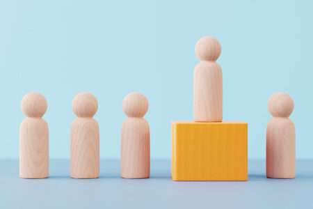 Photo: Wooden figurine pawn standing on wooden cube above other wooden figurines against minimal blue background. The concepts of competition, success, leadership, winning.