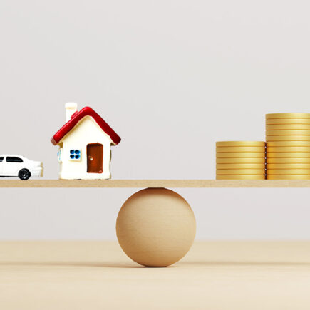 Coins stack on wooden seesaw on the right side, a car and house model on the left, equally balanced.