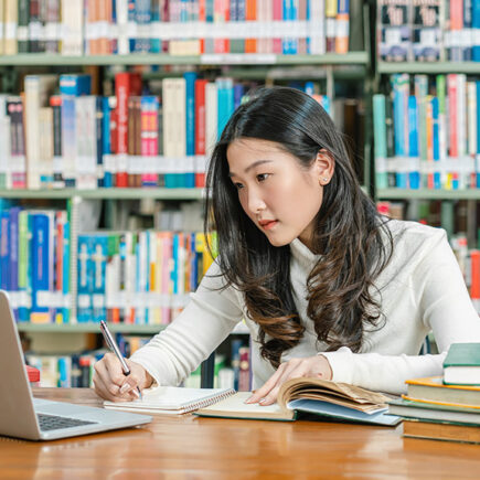 Photo of an Asian female college student in the library in front of shelves of books looking at her laptop and taking notes with books on the desk beside her.