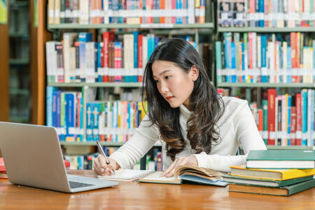 Photo of an Asian female college student in the library in front of shelves of books looking at her laptop and taking notes with books on the desk beside her.