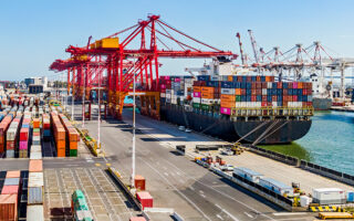 Photo of a port with a large carrier container ship filled up and containers on the pavement near it.