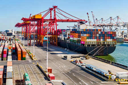 Photo of a port with a large carrier container ship filled up and containers on the pavement near it.