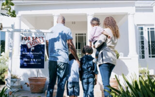 Decorative Image: A young family standing in front of the new home they purchased with the for sale/ sold sign next to them.
