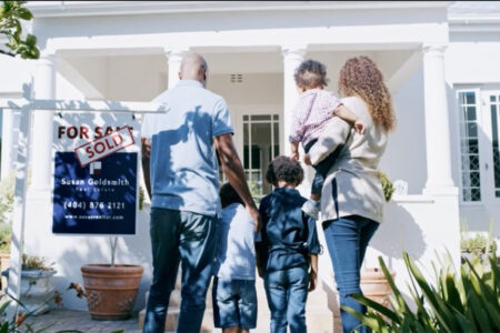 Decorative Image: A young family standing in front of the new home they purchased with the for sale/ sold sign next to them.