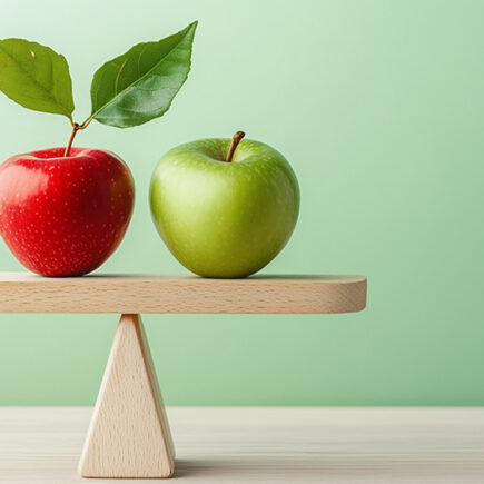 Photo of two apples on a seesaw that is horizontally stable; one is red with two bright green leaves sticking up off the stem; the other is a green apple with stem and no leaves. On a light green background.