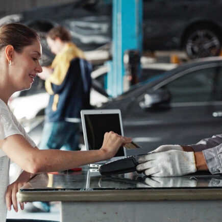 Photo of a car mechanic handing a woman customer a card reader in order to have her pay with credit card. She is placing her credit card on the reader.