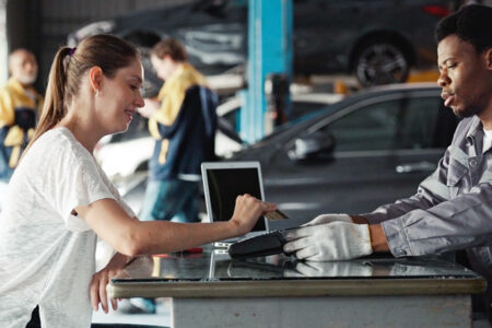 Photo of a car mechanic handing a woman customer a card reader in order to have her pay with credit card. She is placing her credit card on the reader.