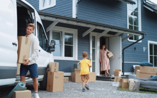Photo: young couple with boy child bringing moving boxes into a newly purchased home that is blue clapboard and white trimmed windows. Boy is bringing his mother, who is standing in the doorway, a plant that looks like lavender.