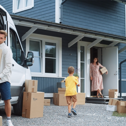 Photo: young couple with boy child bringing moving boxes into a newly purchased home that is blue clapboard and white trimmed windows. Boy is bringing his mother, who is standing in the doorway, a plant that looks like lavender.