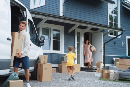 Photo: young couple with boy child bringing moving boxes into a newly purchased home that is blue clapboard and white trimmed windows. Boy is bringing his mother, who is standing in the doorway, a plant that looks like lavender.
