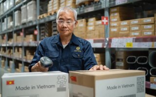 AI generated image of an Asian man in a warehouse with several shelves of cardboard boxes behind him as he scans two boxes in front of him getting ready to ship. Boxes say made in Vietnam.