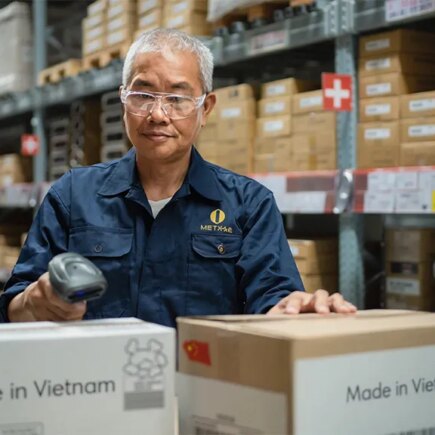 AI generated image of an Asian man in a warehouse with several shelves of cardboard boxes behind him as he scans two boxes in front of him getting ready to ship. Boxes say made in Vietnam.