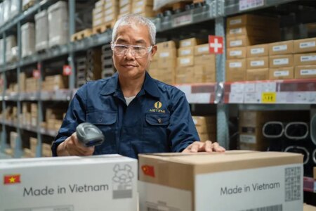 AI generated image of an Asian man in a warehouse with several shelves of cardboard boxes behind him as he scans two boxes in front of him getting ready to ship. Boxes say made in Vietnam.