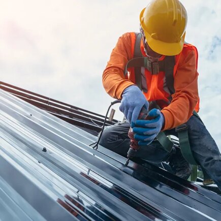Roofer worker in special protective work wear and gloves,using air or pneumatic nail gun and installing asphalt or bitumen shingle on top of the Metal Sheet roof.