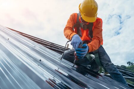 Roofer worker in special protective work wear and gloves,using air or pneumatic nail gun and installing asphalt or bitumen shingle on top of the Metal Sheet roof.