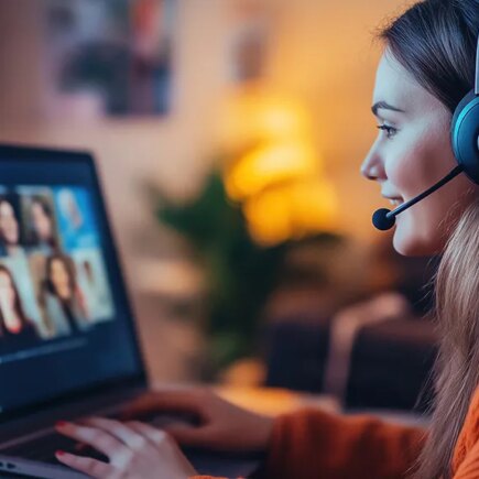 Photo of young caucasian woman working at her desk at home wearing a headsset and engaged in a videoteleconferencing call on her laptop computer.
