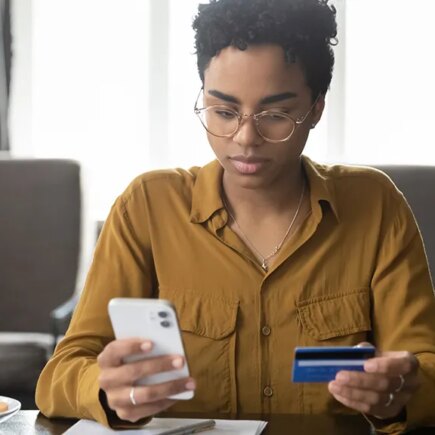 Serious young Black bank customer woman paying for purchase, domestic fees, bills by blue plastic credit card, using online payment app on smartphone, shopping on internet stores