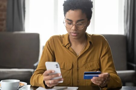Serious young Black bank customer woman paying for purchase, domestic fees, bills by blue plastic credit card, using online payment app on smartphone, shopping on internet stores