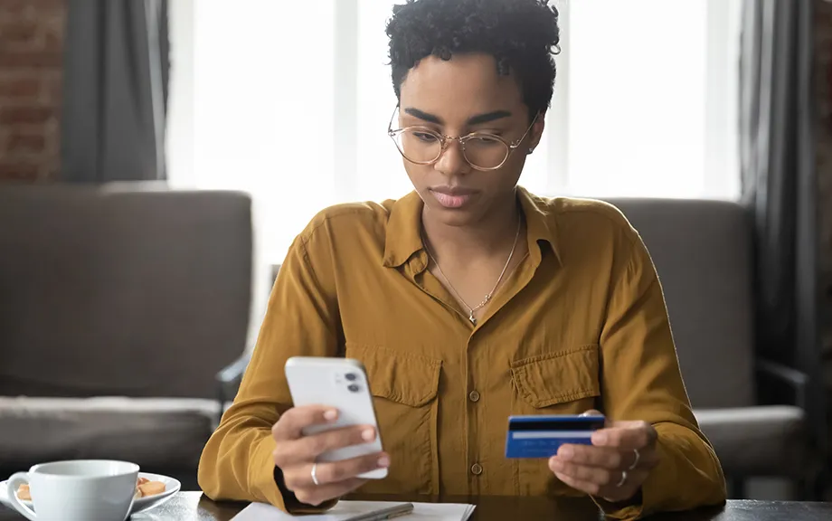 Serious young Black bank customer woman paying for purchase, domestic fees, bills by blue plastic credit card, using online payment app on smartphone, shopping on internet stores